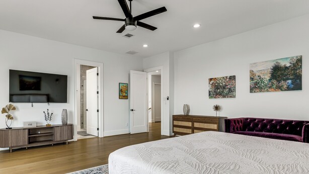Un dormitorio con cama, ventilador de techo, televisor, cómoda y obras de arte en las paredes.