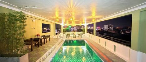 Una piscina en la azotea con suelo a cuadros, rodeada de asientos y mesas, con vista a un paisaje urbano nocturno.