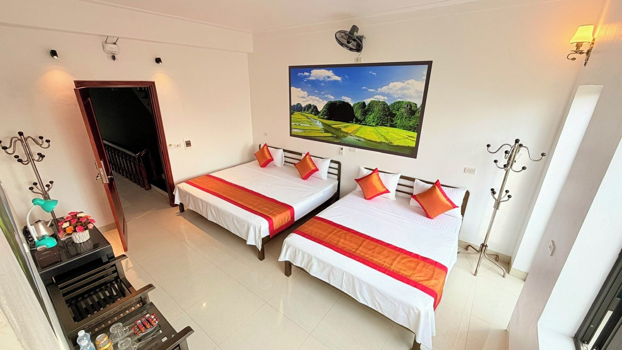 Habitación de hotel con dos camas, un cuadro de un paisaje, una mesita con una lámpara y un perchero.