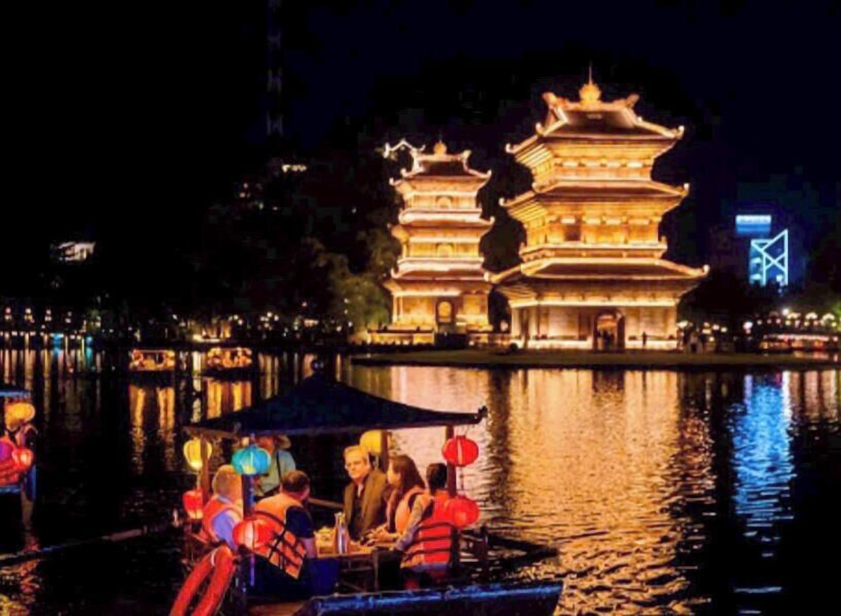 Une pagode traditionnelle à plusieurs niveaux, illuminée la nuit, dont le reflet se mire sur une étendue d’eau calme.
