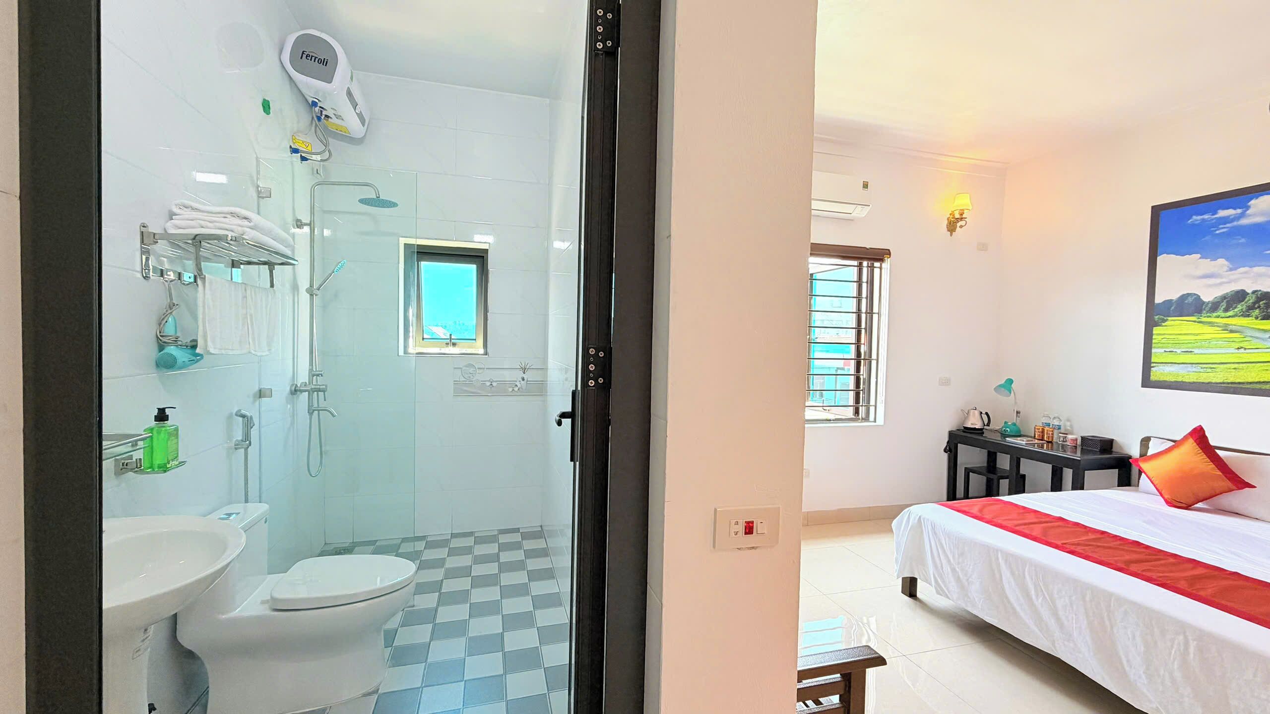 Habitación de hotel con baño, cama con almohada roja decorativa, pintura de un paisaje y ventana con vista a edificios.
