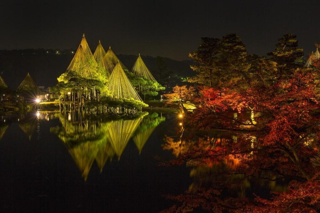 A nighttime view of a traditional structure with multiple conical roofs, surrounded by a body of water and trees with autumn foliage.