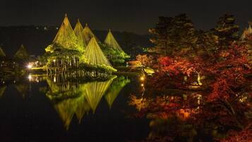 A nighttime view of a traditional structure with multiple conical roofs, surrounded by a body of water and trees with autumn foliage.