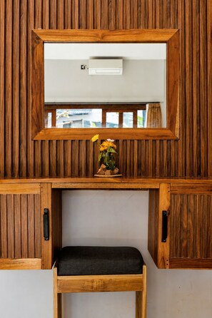 Un miroir à cadre en bois est placé au-dessus d’un banc agrémenté d’un coussin, et une petite plante aux fleurs jaunes se trouve sur une étagère en dessous.