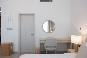 A hotel room with a bed, a desk with a chair, a round mirror, a bedside lamp, and a trash bin.