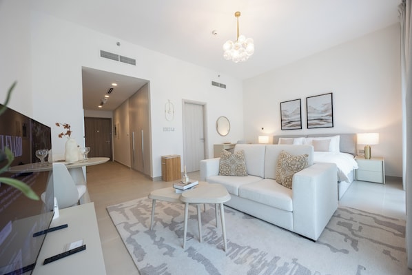 A modern living room with a sofa, a coffee table, and a TV. There is a hallway leading to a bedroom with a bed and a nightstand.