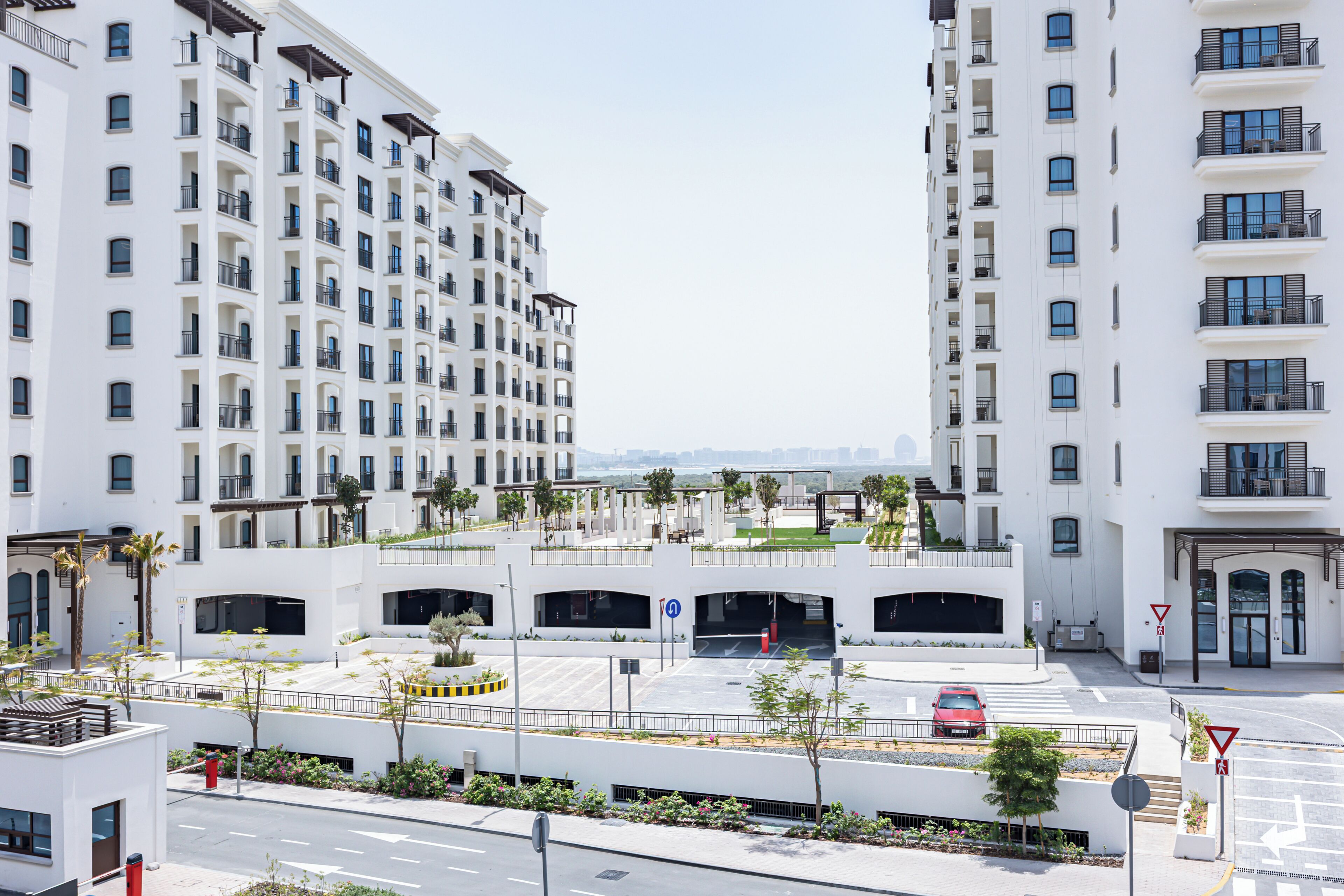 A modern multi-story building with balconies, a parking area, and a pedestrian walkway.