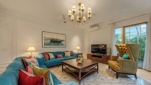Una sala de estar con un sofá azul, una mesa de centro de madera, un sillón dorado, un televisor y un ventanal con vistas a la vegetación.