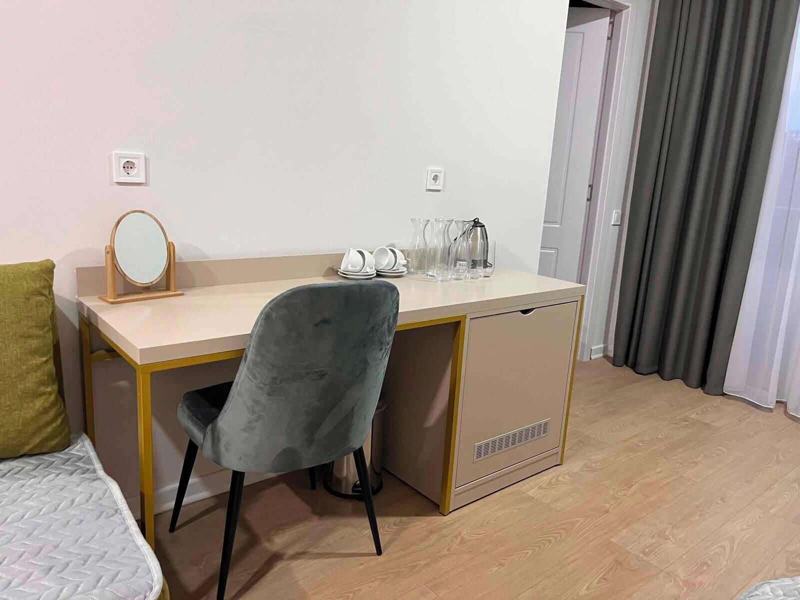 Ein Hotelzimmer mit Bett, Schreibtisch mit Spiegel, Stuhl, einer kleinen Küchenzeile und einem Fenster mit Vorhängen.
