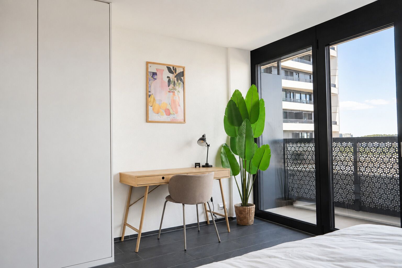 Une chambre moderne avec un lit, un bureau et une chaise, une grande plante verte et une fenêtre donnant sur des immeubles.