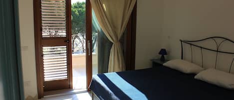 Une chambre à coucher comprenant un lit, une table de chevet, une lampe, une fenêtre avec des rideaux et une porte donnant sur un espace extérieur.