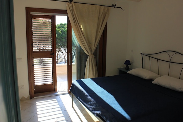 A bedroom with a bed, a nightstand, a lamp, a window with curtains, and a door leading to an outdoor area.