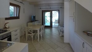 Una cocina con muebles blancos, una mesa de comedor con sillas, un televisor sobre un mueble bajo y vistas a un área exterior a través de un ventanal.