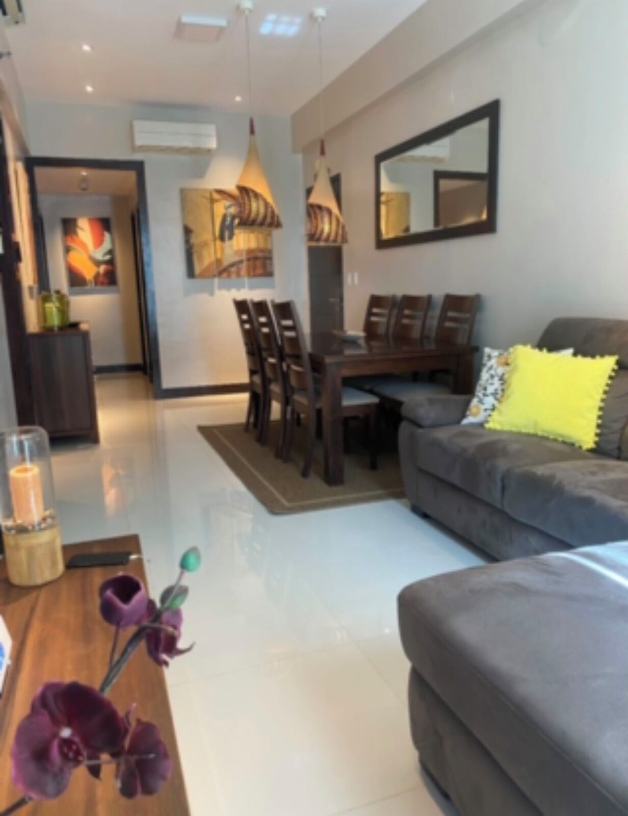 Uma sala de estar moderna com um sofá cinzento, uma mesa de jantar com cadeiras e uma mesa de centro com um vaso de flores.