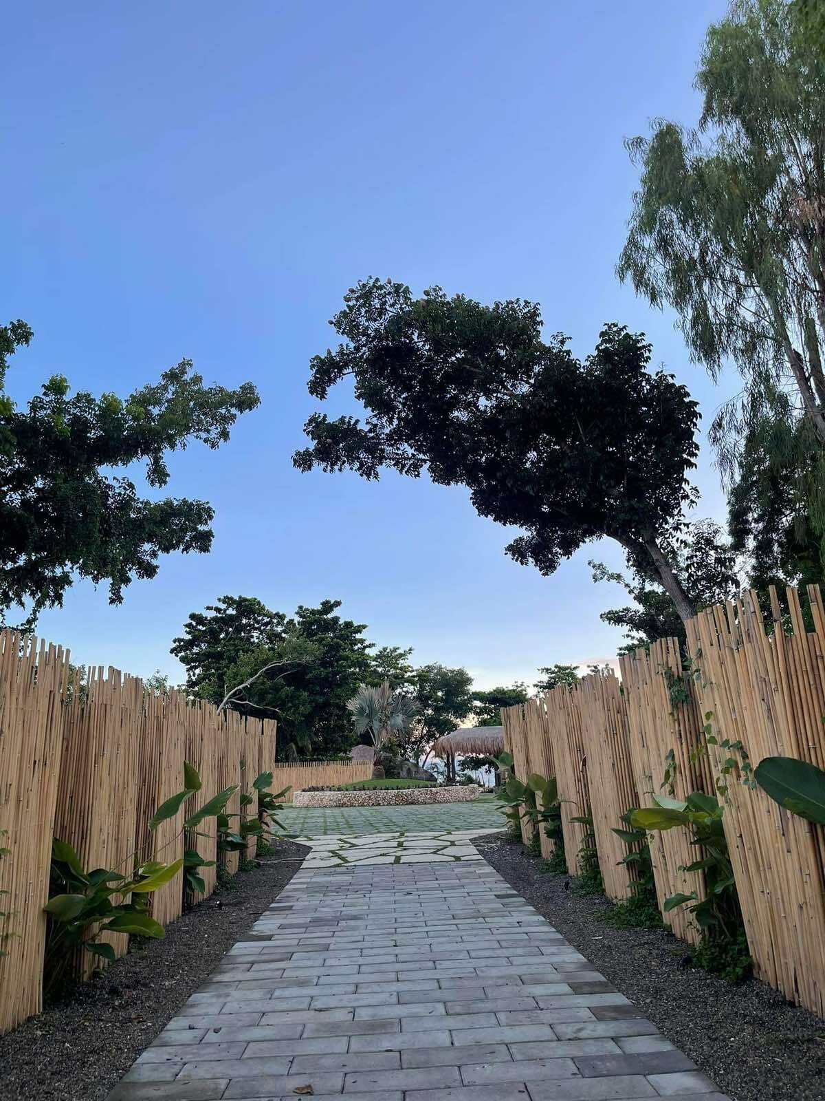 Un chemin pavé, bordé de clôtures en bois, mène à un espace jardin agrémenté d’arbres et surmonté d’une structure à toiture de chaume.
