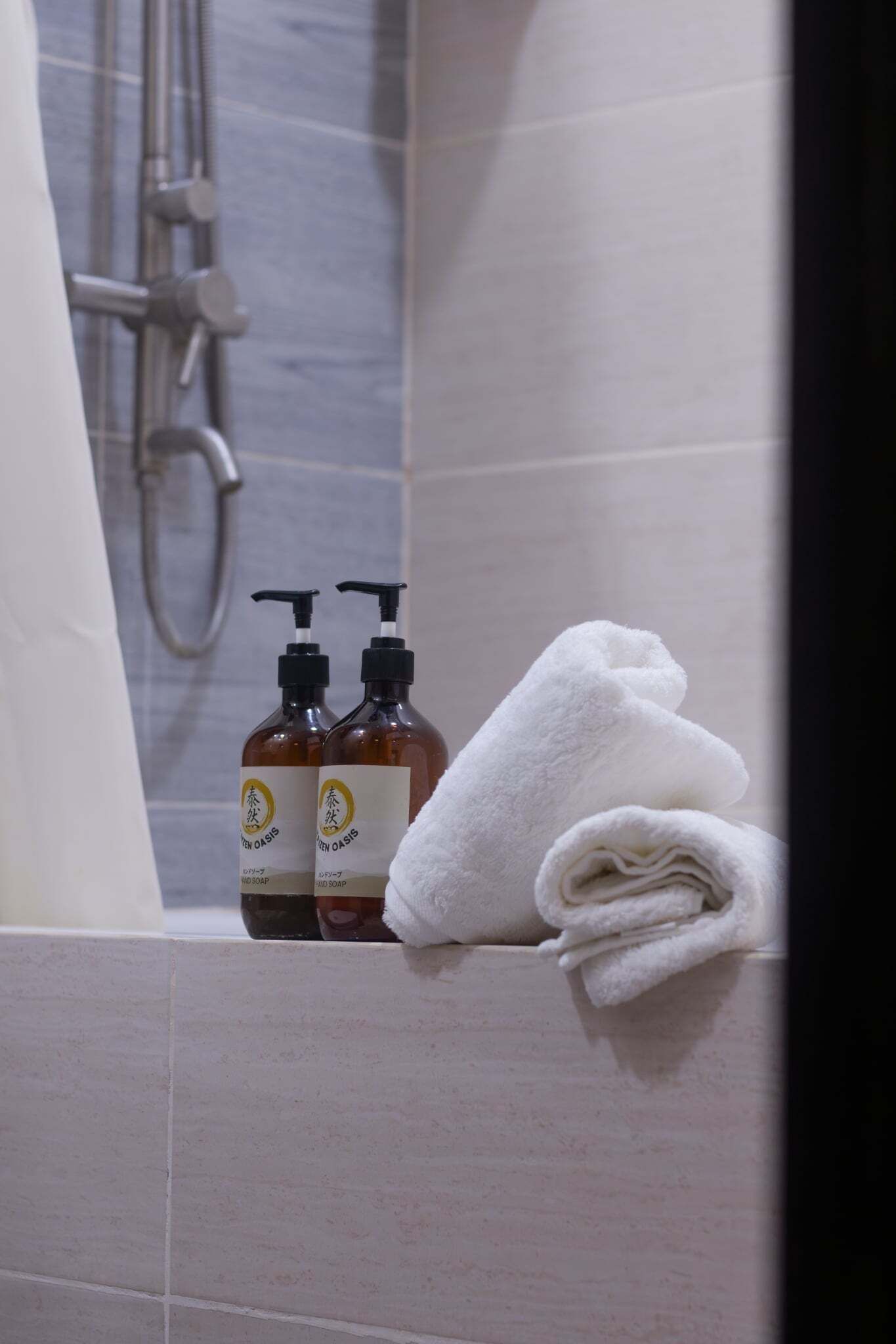 Une salle de bain avec une serviette blanche enroulée sur une étagère, deux bouteilles de savon liquide et une pomme de douche.