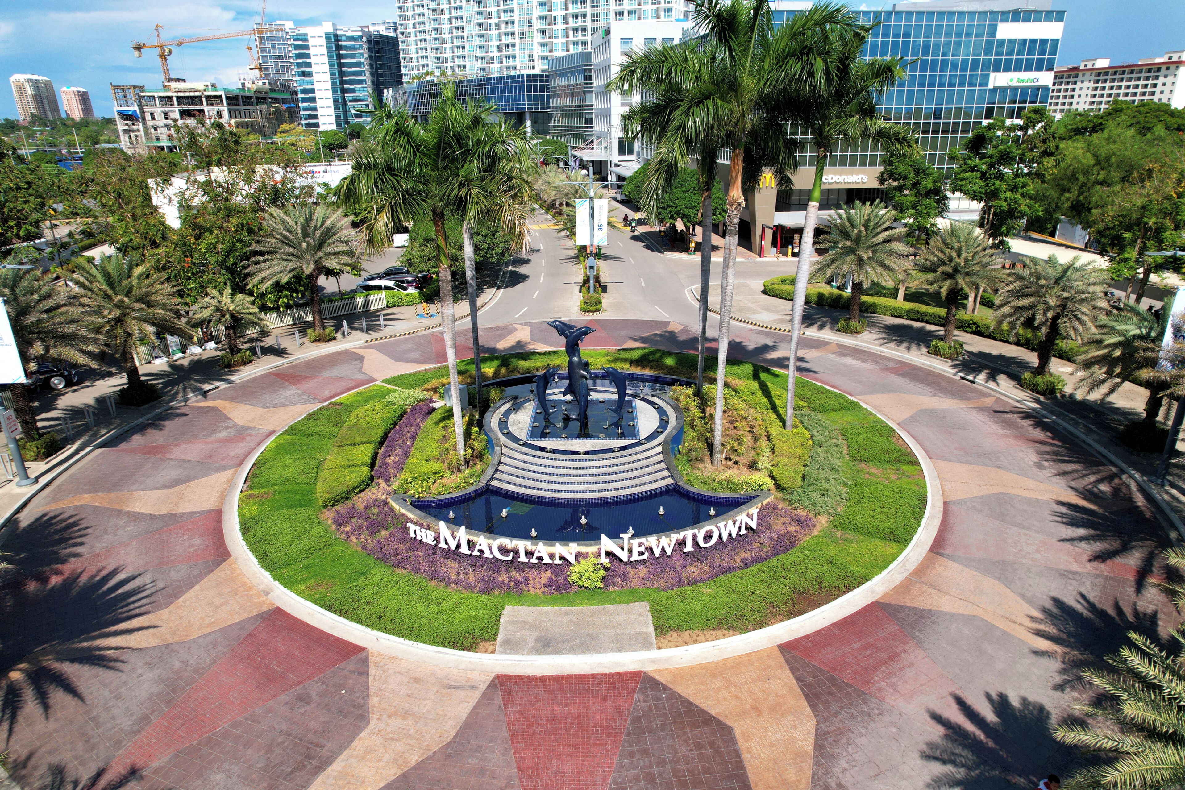 A circular plaza with a central fountain, surrounded by palm trees and greenery, with modern buildings in the background.