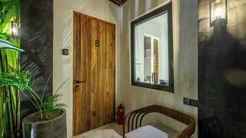 Una puerta de madera con un número, una planta en maceta, una silla y un espejo montado en la pared.