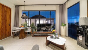 A modern living room with a dining area, a large TV, and a view of a building through the windows. - Sebelas Apartments Ubud (Gianyar)
