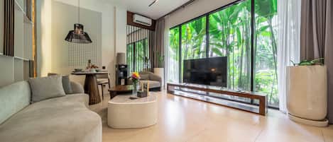 A modern living room with a large TV, comfortable seating, a coffee table, and a view of greenery through large windows.