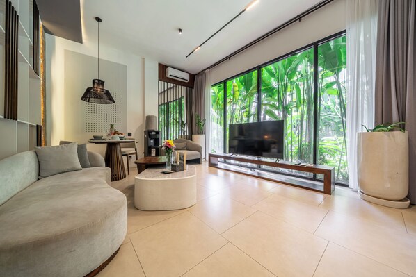 A modern living room with a large TV, comfortable seating, a coffee table, and a view of greenery through large windows. - Sebelas Apartments Ubud (Gianyar)
