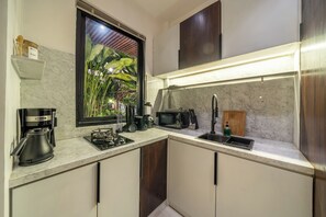 A modern kitchen with a built-in stove, a coffee maker, a sink, and a window with a view of greenery. - Sebelas Apartments Ubud (Gianyar)