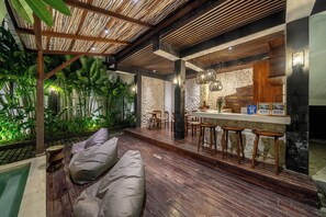 A modern interior with a wooden floor, a seating area with cushions, a bar with stools, and a stone wall. - Sebelas Apartments Ubud (Gianyar)