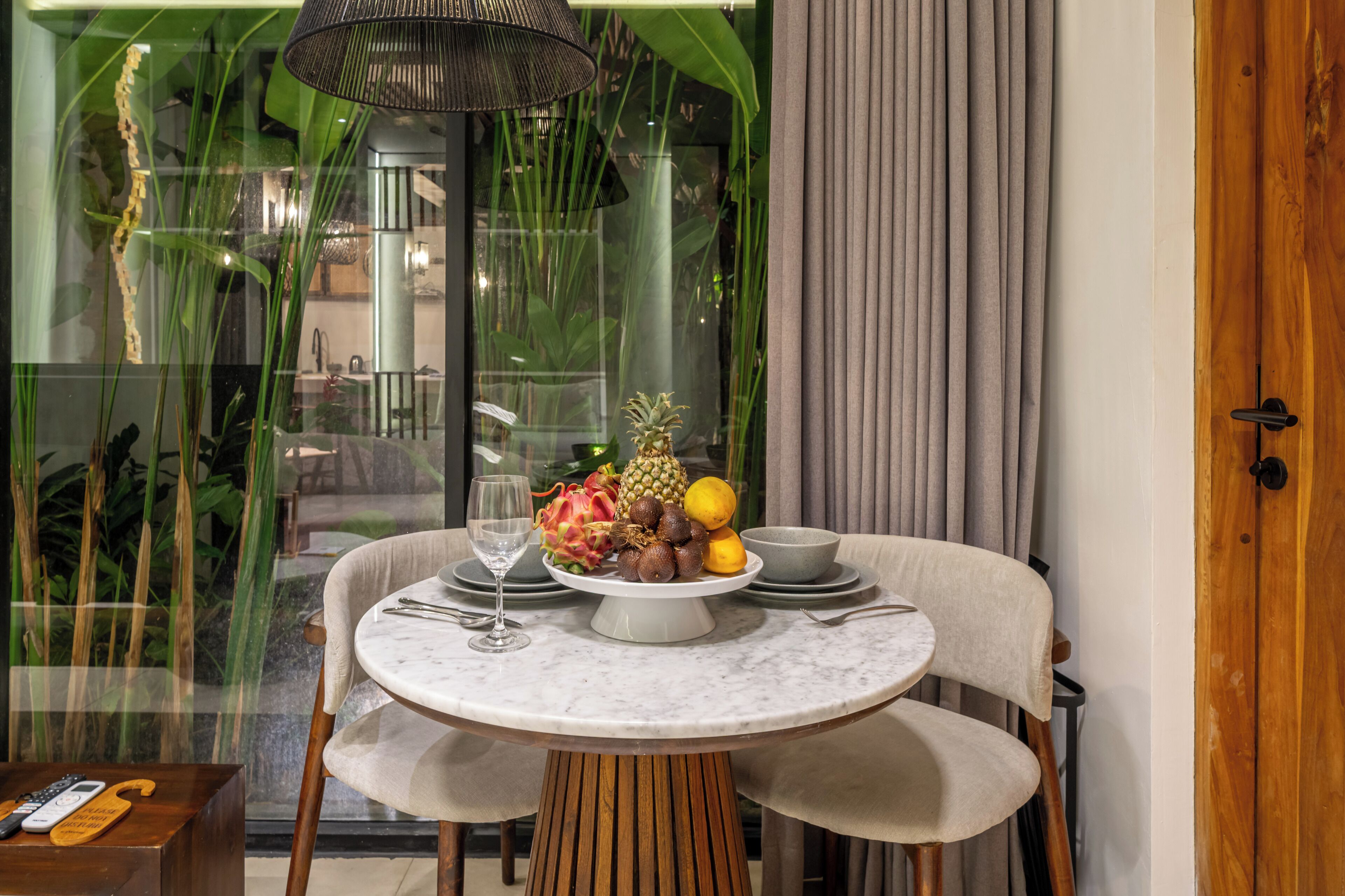 Una mesa de comedor con frutero, copa de vino y cubiertos, ubicada en un espacio con vista a follaje exterior.