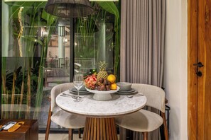 A dining table with a fruit bowl, a wine glass, and cutlery, set in a room with a view of greenery outside. - Sebelas Apartments Ubud (Gianyar)