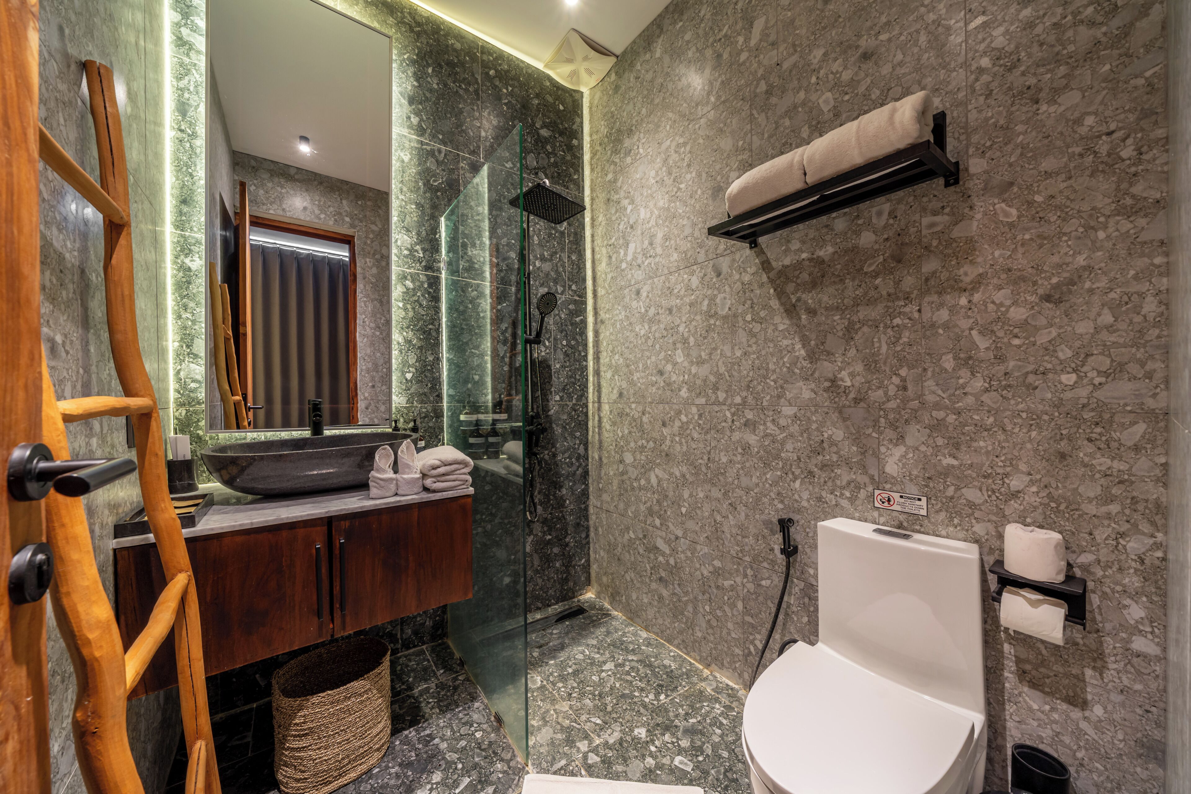 Een badkamer met een marmeren wand, een houten wastafelmeubel, een douchecabine van glas en een toilet.
