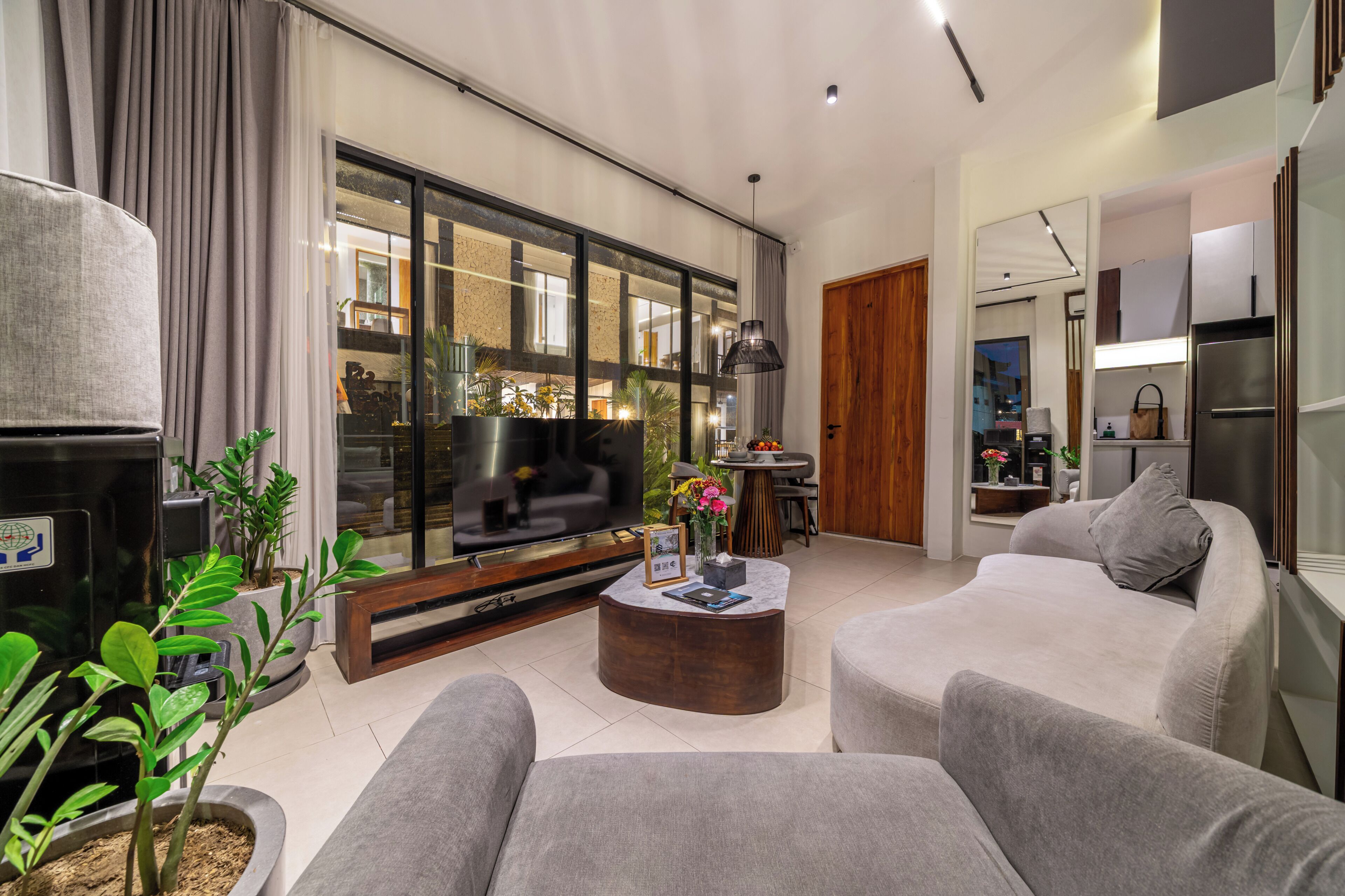 A modern living room with a large flat-screen TV, grey sofas, a coffee table, and a dining area with a wooden door.