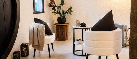 A modern living room with a large wooden door, a round table, two chairs, a potted plant, and a wall clock.