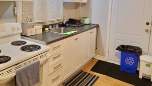 Een kleine keuken met witte kastjes, een fornuis, een spoelbak en een prullenbak.