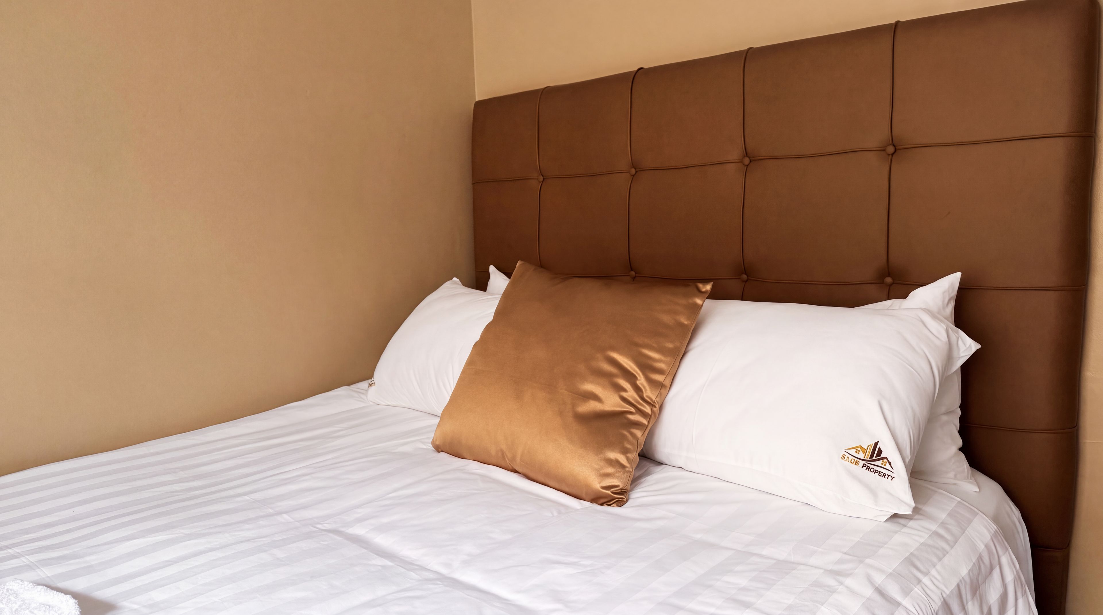 Una cama con cabecera capitonada, ropa de cama blanca y una sola almohada marrón.