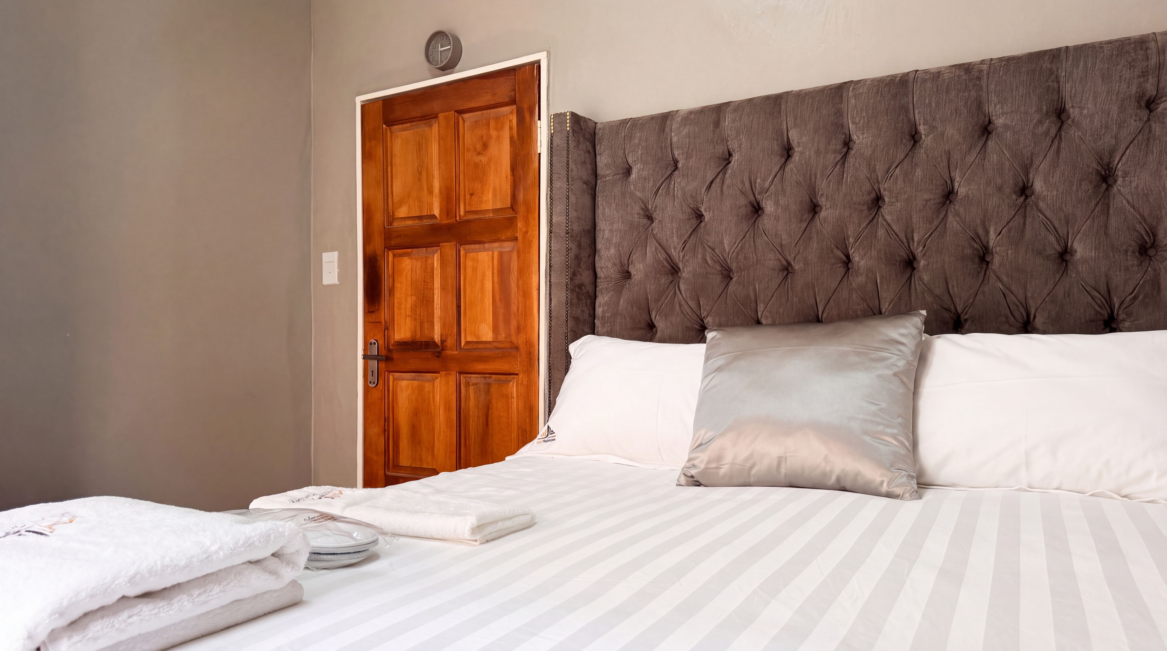 Una cama bien hecha con ropa de cama a rayas y dos almohadas, una puerta de madera con un reloj, e interruptor de luz fijado a la pared.