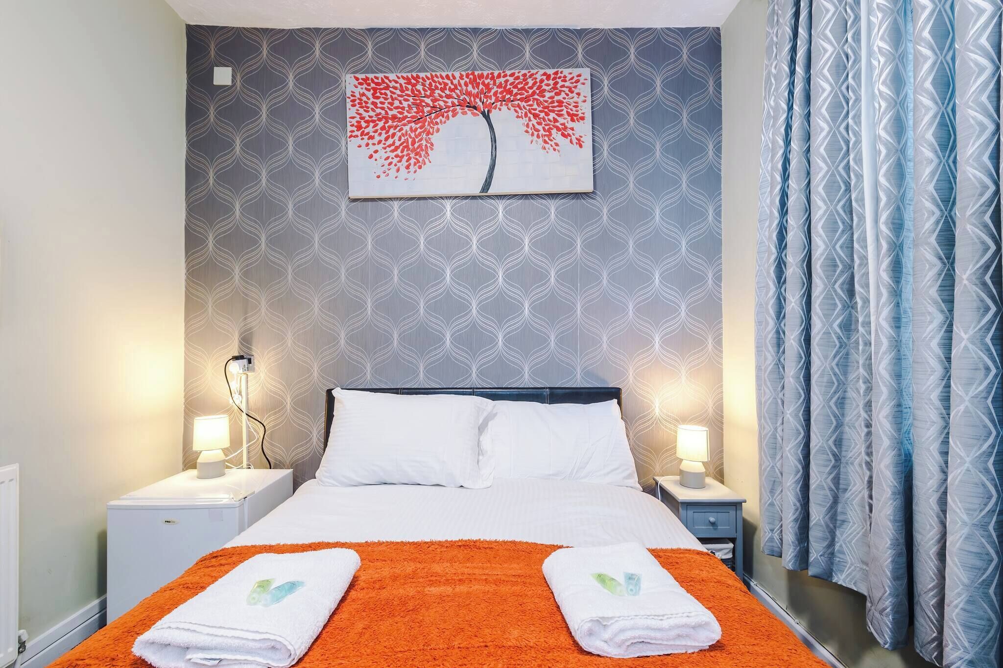 A hotel room with a bed, two bedside tables, a nightstand, a wall-mounted painting, and a patterned curtain.