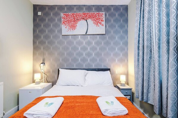 A hotel room with a bed, two bedside tables, a nightstand, a wall-mounted painting, and a patterned curtain.