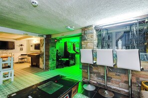 A room with a green-lit gaming area, a fire exit sign, a television, and a bar area with stools.