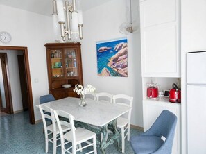 A dining area with a marble table, white chairs, a cabinet with glass doors, and a painting of a coastal scene. - Modern Apartment Near Senigallia's Velvet Beach (Senigallia)
