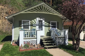 Ein kleines, eingeschossiges Haus mit Veranda, ein kleiner Garten mit Steinen und wenigen roten Blumen, sowie ein Baum mit rosa Blüten.