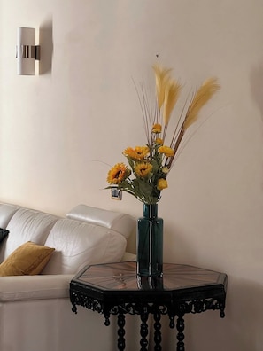 Um quarto com um sofá branco, um vaso verde-água com flores amarelas e uma mesa preta com tampo de vidro.