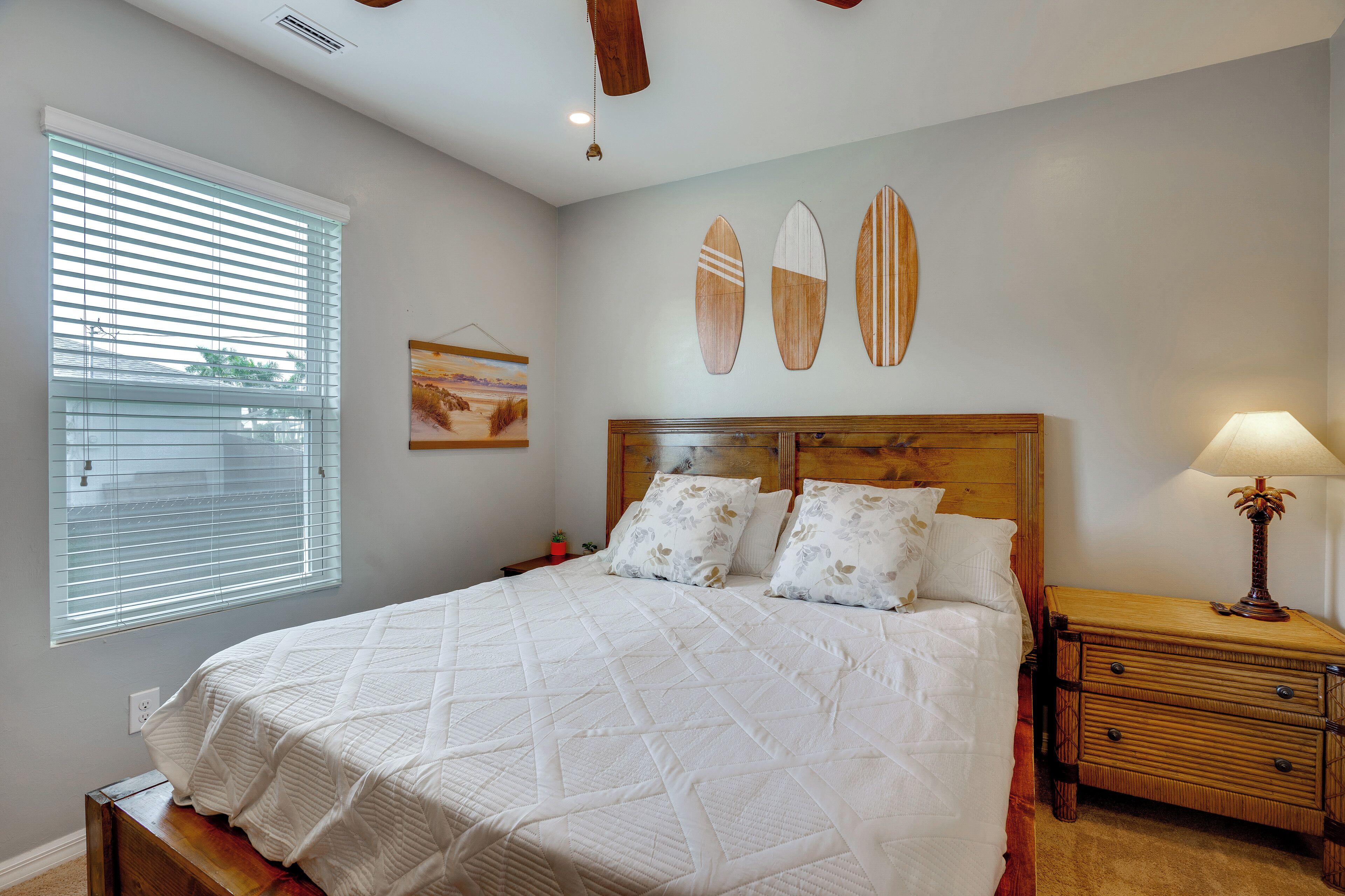A bedroom with a bed, a nightstand, a lamp, a window with blinds, and a wall with three surfboards.