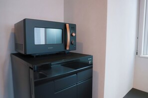 A microwave oven on top of a black appliance, possibly a coffee maker or a small refrigerator, in a room with a plain wall and a window with a curtain.
