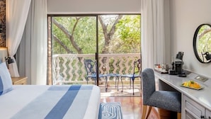 A hotel room with a bed, a desk with a coffee maker, a chair, and a balcony with a view of trees.