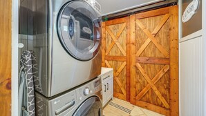 A laundry area with stacked washing machines, a wooden door, and a sign indicating self-service. - Swings, Fire Pit + Views: Shirleysburg Cabin! (Shirleysburg)