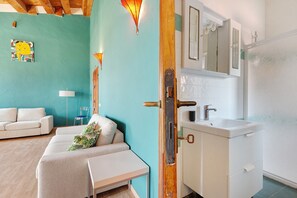 Habitación con pared color aguamarina, un sofá blanco, una mesita, una silla transparente, una lámpara y un baño con lavamanos y espejo blancos.