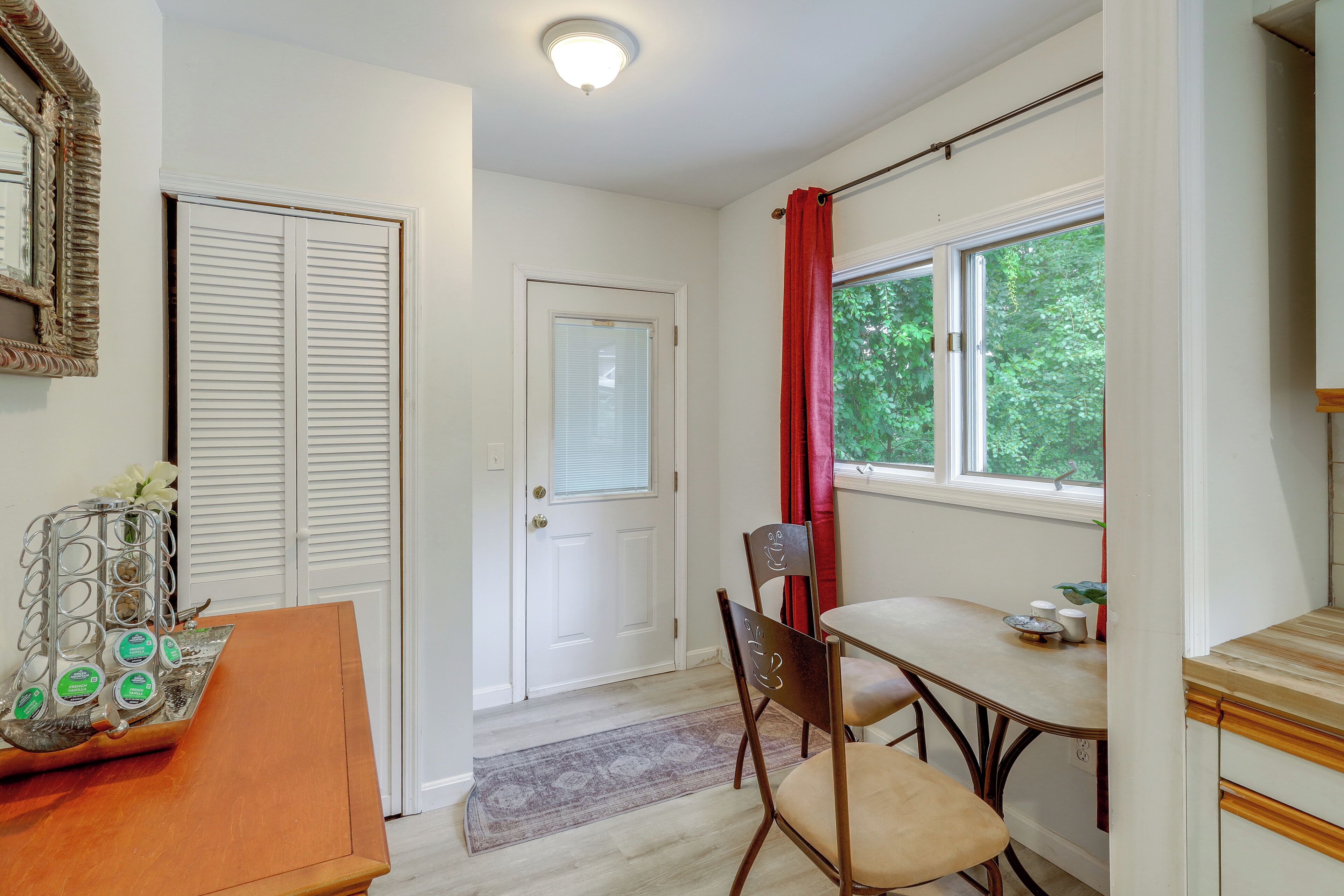 A room with a white door, a window with red curtains, a table with chairs, and a mirror with an ornate frame.