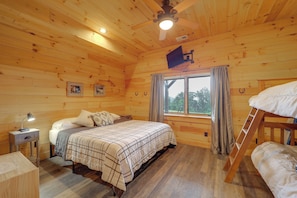 Un dormitorio de cabaña de madera con cama, escritorio, ventilador de techo y ventana con cortinas.