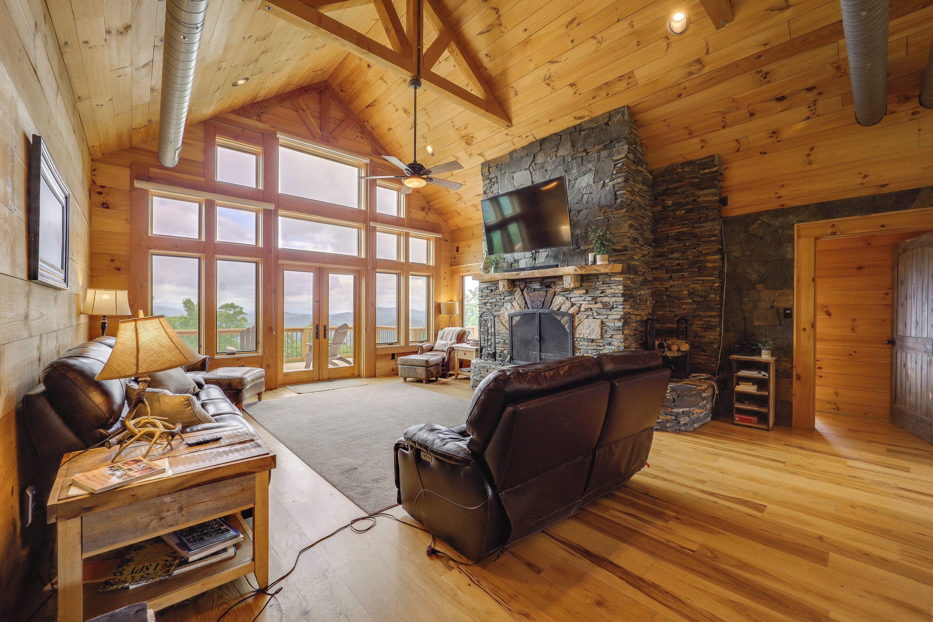 A spacious living room with a stone fireplace, leather sofas, and a large TV mounted on the wall.