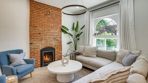 Uma sala de estar com lareira de tijolos, um sofá, uma poltrona azul e uma mesa de centro redonda.